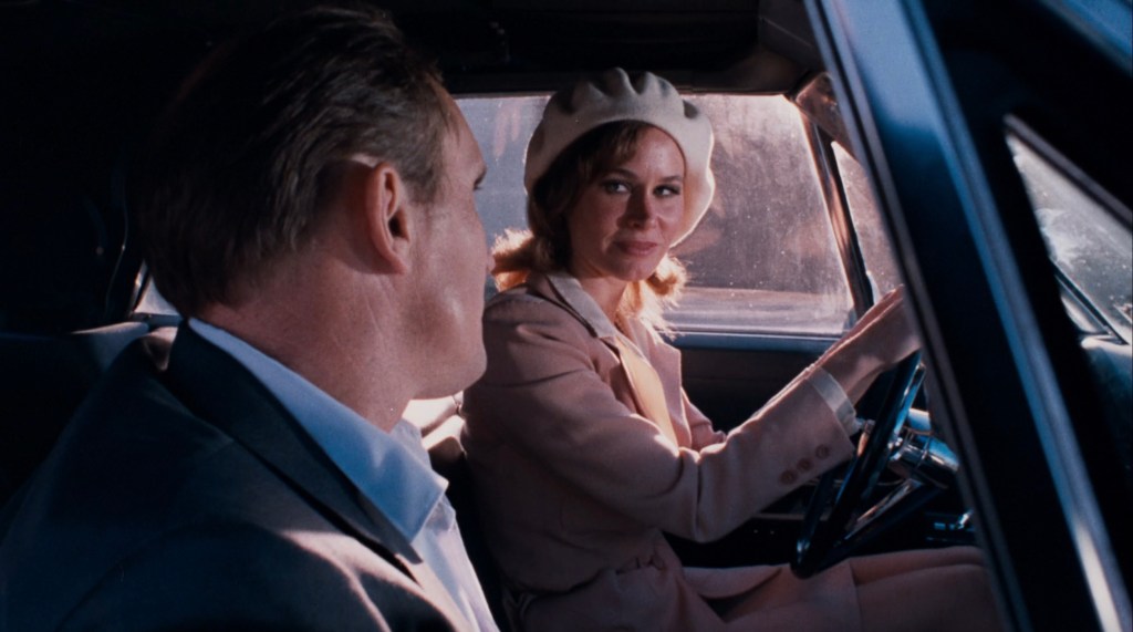 A woman wearing a beret and a light pink outfit smiles while sitting in the driver's seat of a car, facing a man who looks at her.