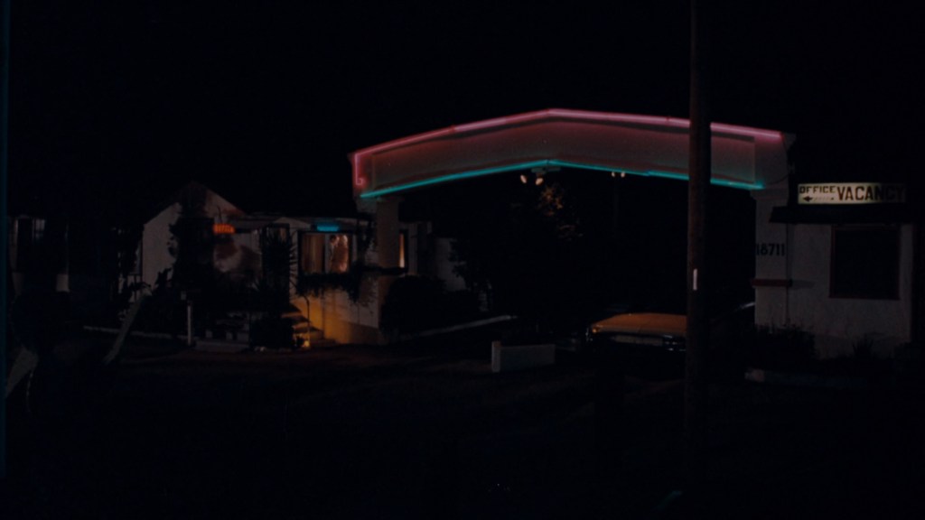 A motel at night with neon lights displaying 'VACANCY' and an illuminated office sign. The scene captures the entrance and part of the building, highlighting a retro ambiance.