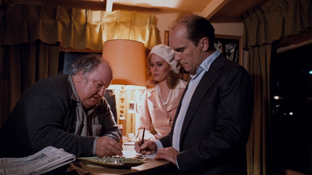 A scene depicting three individuals in an indoor setting, with two men and one woman engaged in conversation around a table, illuminated by a lamp. One man is writing while the other observes, and the woman stands between them, looking on.