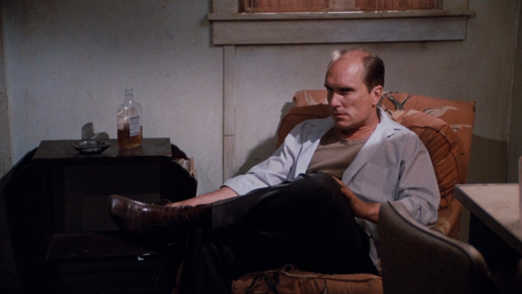 A man sitting in an armchair in a dimly lit room, dressed in a light blue jacket over a beige shirt, with a serious expression on his face. A bottle on a table nearby suggests a casual, lived-in environment.