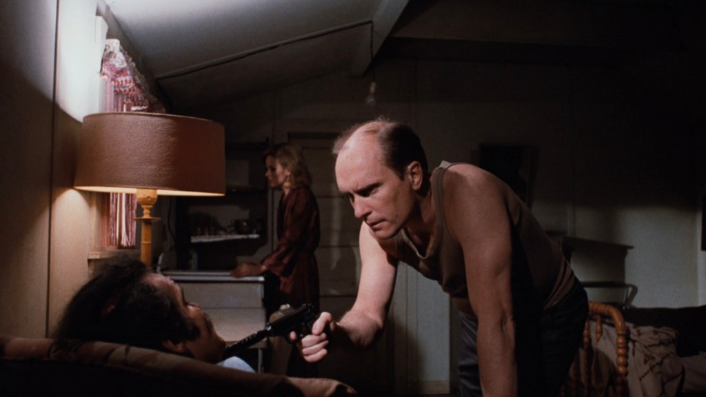 A tense scene featuring a man in a tank top pointing a gun at another man seated on a couch, while a woman stands in the background.