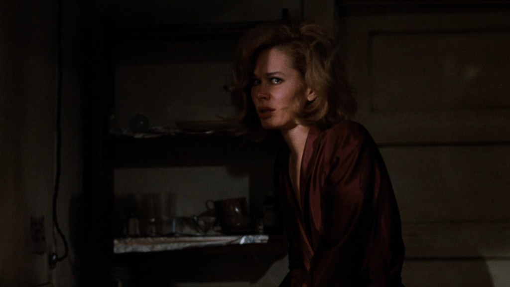 A woman with disheveled hair sits in a dimly lit room, wearing a dark robe, looking apprehensive or distressed.