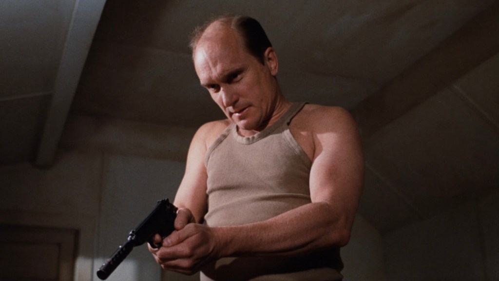 A man in a tank top holding a handgun, looking intently at the weapon.