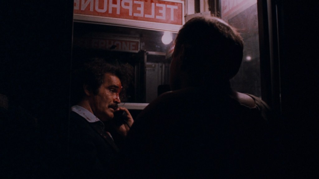 A man with a mustache is talking on the phone inside a phone booth, showing signs of distress, while another person stands outside the booth, partially visible.