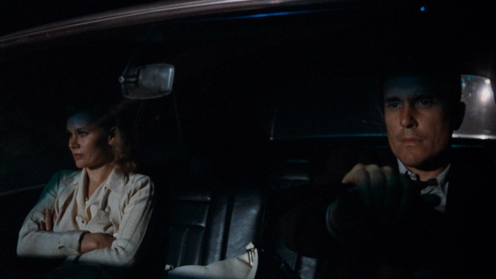 A man driving a car with a serious expression, while a woman sits beside him, looking troubled and arms crossed.