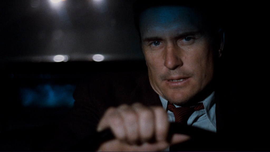 A close-up of a man driving at night, showing a serious expression and focused demeanor, with dim lighting and headlights visible in the background.