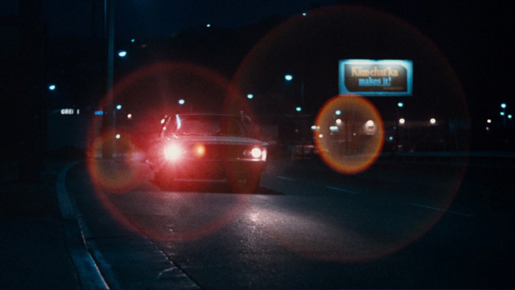 A car with its headlights on driving down a dimly lit street at night, with a billboard advertising 'Kimchuka' in the background.