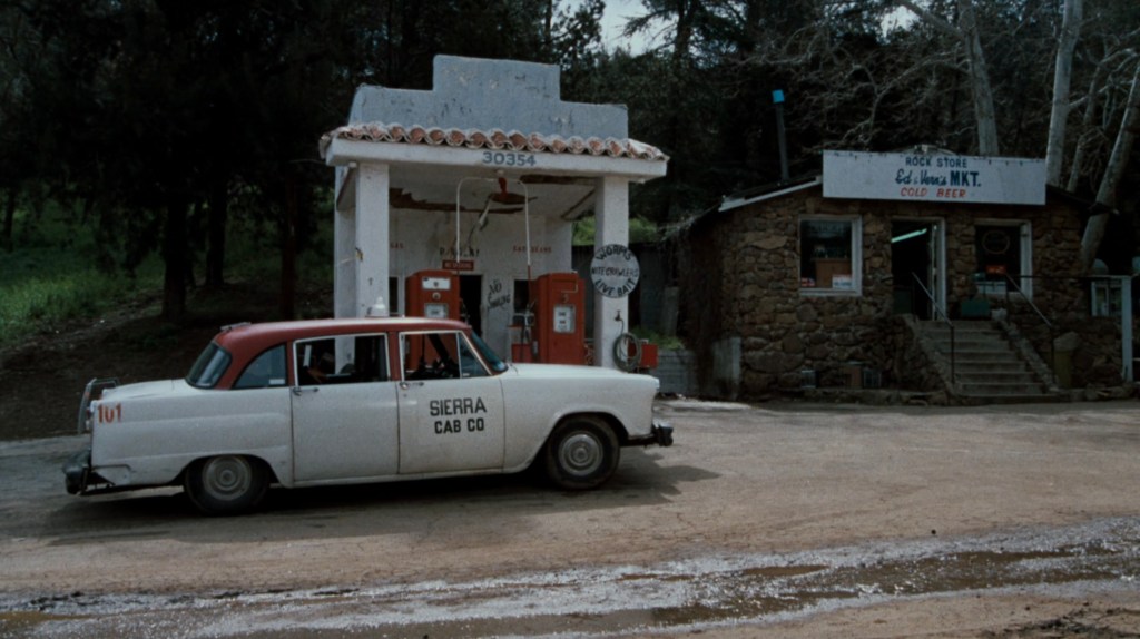 A vintage cab labeled 'Sierra Cab Co.' parked at a small gas station with red fuel pumps in a rural setting. Nearby, there is a stone building with a sign for a rock store.