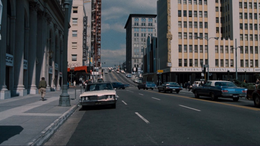 A street scene featuring vintage cars parked along the roadside and a city sidewalk. Buildings line both sides of the street, with a visible slope and a mix of architecture. People can be seen walking in the distance under a partly cloudy sky.