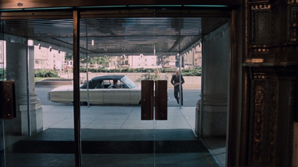 View from inside a building through glass doors, showing a man walking outside towards a classic white car parked on the street.