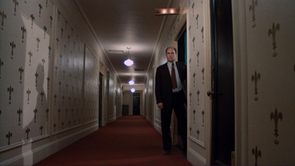 A man in a suit stands in a hotel corridor with patterned wallpaper and red carpeting, partially illuminated by overhead lights.
