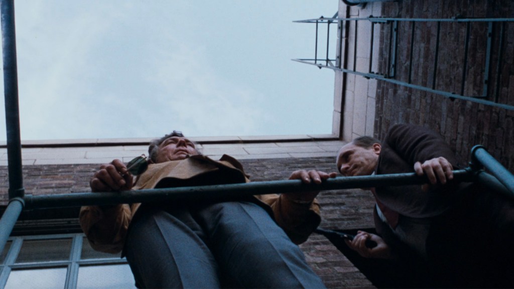 Two men are seen from below, looking up at a building while holding onto a railing. One man is wearing a brown jacket and the other is in a dark coat, creating a tense moment with a sense of urgency.