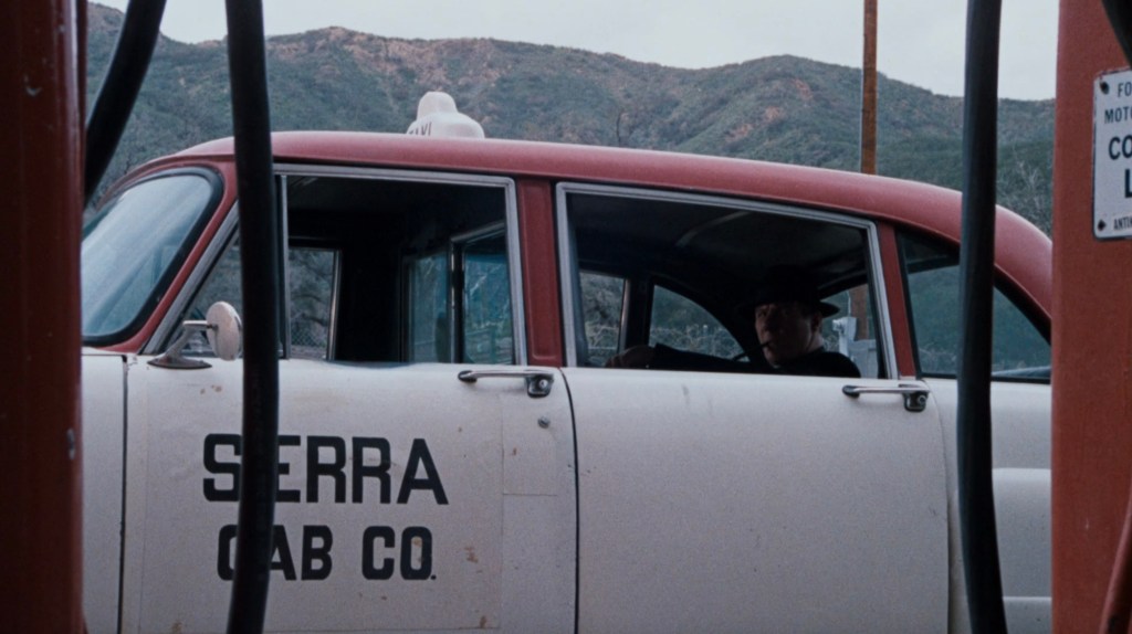 A vintage taxi cab with 'Sierra Cab Co' written on the side, parked at a gas station with mountains in the background. A person wearing a hat is sitting in the driver's seat.