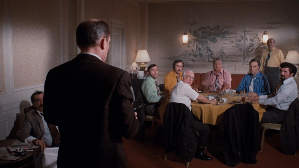 A group of men sitting around a table in a meeting room, with one man standing and speaking to them. The walls are adorned with a mural, and the men appear to be engaged in a serious discussion.