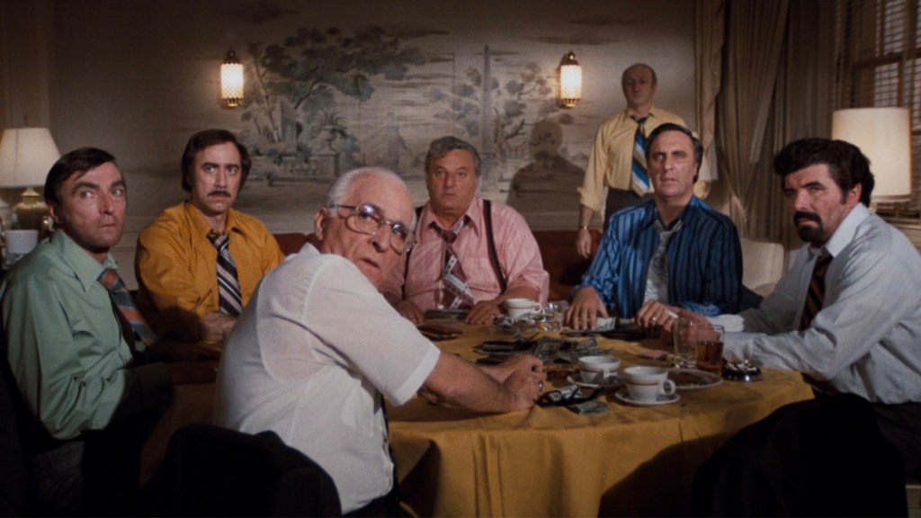 A group of seven men seated around a table, looking directly at the camera. The men are dressed in various colorful shirts and ties, with an array of coffee cups and playing cards scattered on the table.