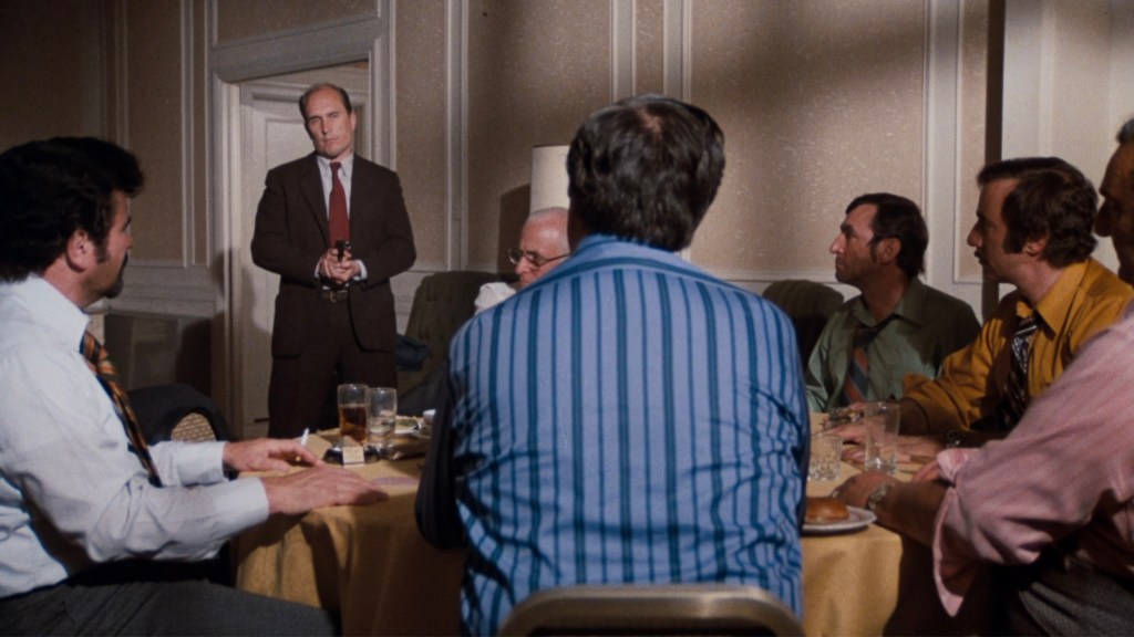 A group of men seated around a table in a dimly lit room, with one man standing and speaking, holding a gun. The men are dressed in formal attire, and the table has drinks and food.