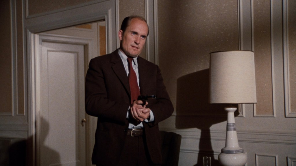 A man in a brown suit holding a gun in a well-lit room with a lamp and a doorway in the background.