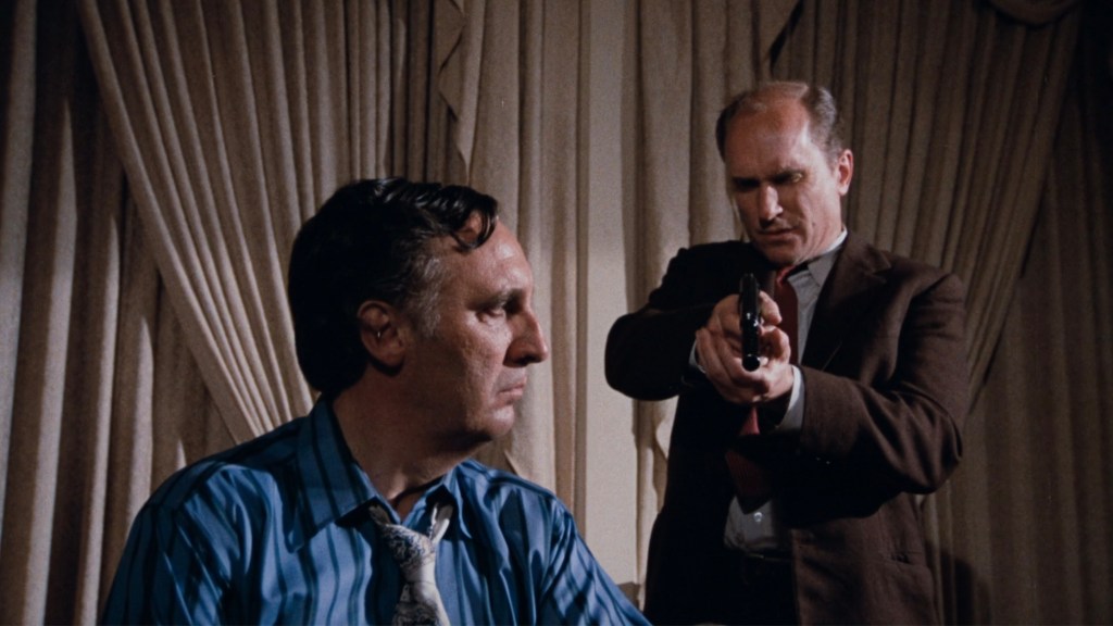 A tense scene featuring a man sitting in a striped shirt with a serious expression, while another man in a suit points a gun at him.