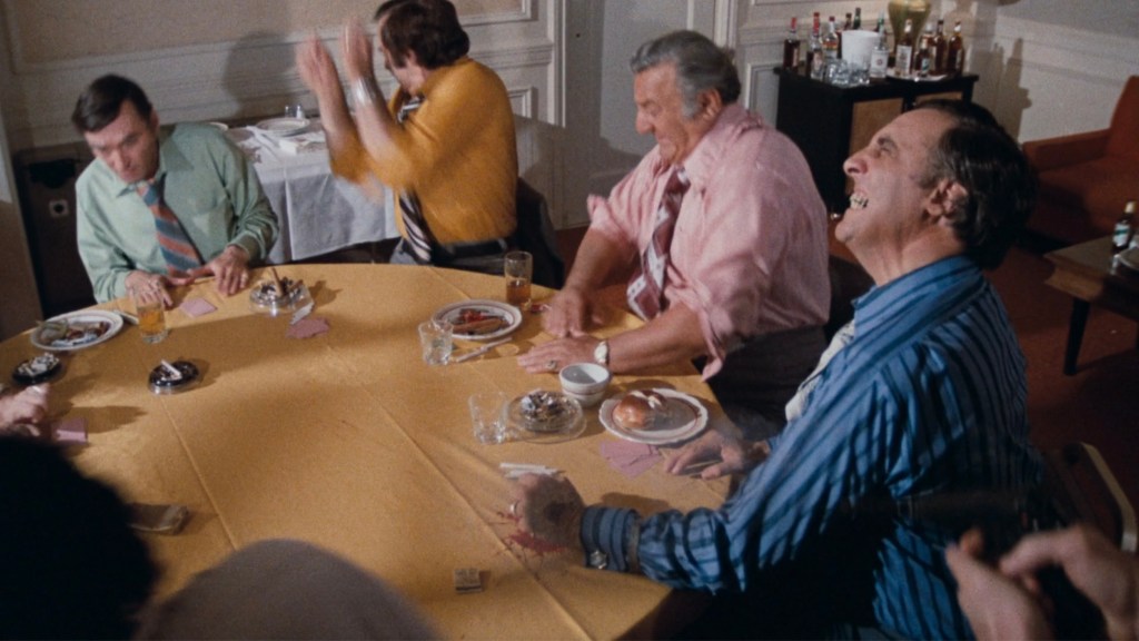 A group of men gathered around a yellow table, engaging in animated conversation with laughter, food, and drinks in a restaurant setting.