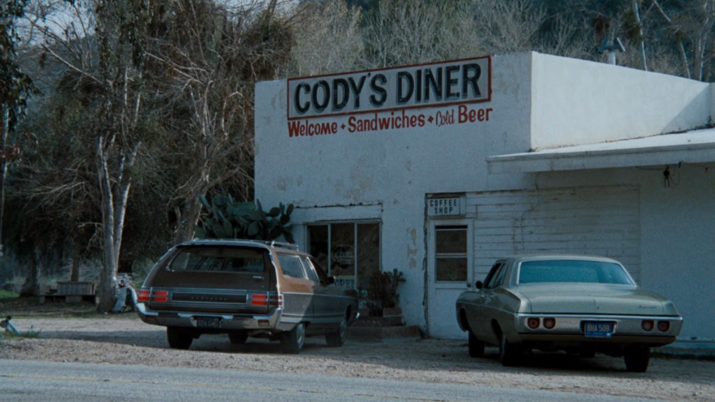 A vintage diner with a sign reading 'Cody's Diner' featuring the words 'Welcome - Sandwiches - Cold Beer.' Two classic cars are parked outside, surrounded by trees.