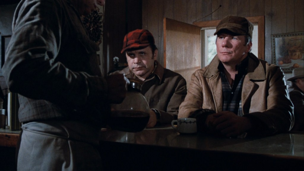 Two men sitting at a bar counter, engaged in conversation while a server pours coffee from a carafe. The setting appears rustic with wooden walls and other patrons in the background.