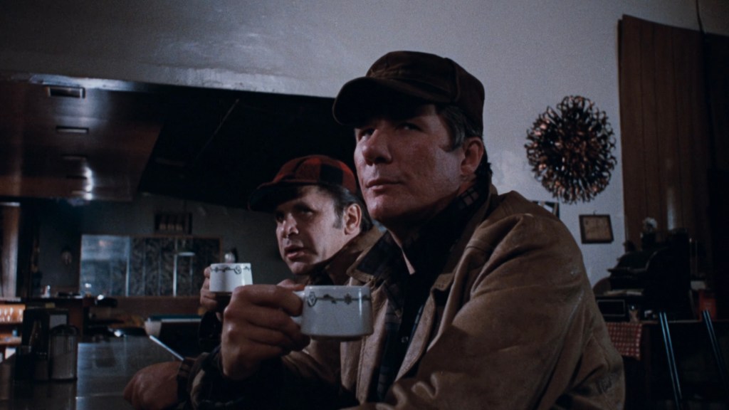 Two men sitting at a bar, one holding a cup while the other gazes intently off to the side. The interior features a retro design with a dark bar and some decor in the background.