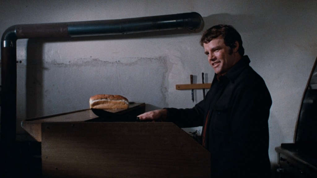 A man standing in a dimly lit room with a smile, positioned near a wooden table with a hot dog bun on it.