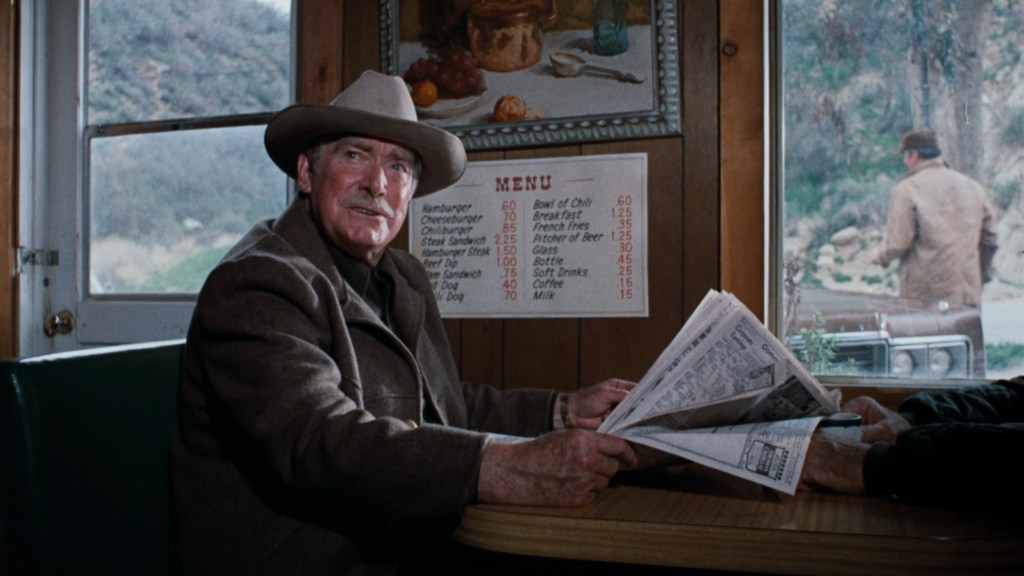 An elderly man wearing a hat sits at a booth in a diner, holding a newspaper and looking towards the camera. A menu is displayed on the wall behind him, and a person can be seen walking outside the diner.