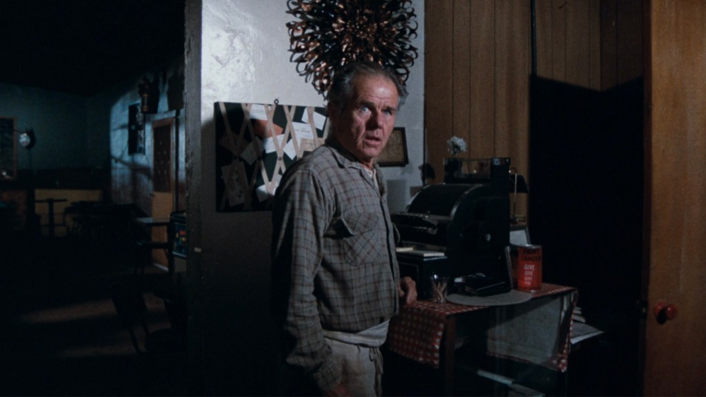 An older man stands in a dimly lit room, looking surprised or concerned. He is wearing a plaid shirt and appears to be near a cash register and a table with a checkered tablecloth.
