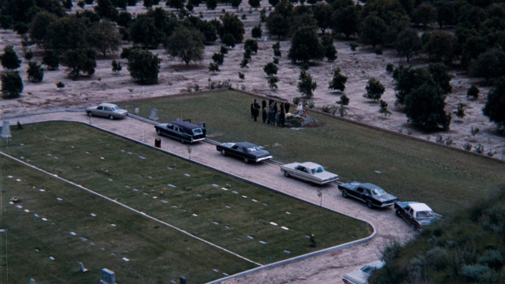 An aerial view of a cemetery with several parked cars and a group of people gathered at a gravesite.