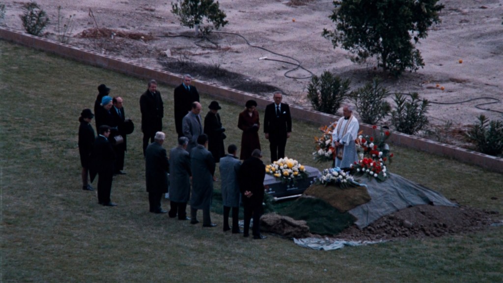A group of people gathered for a funeral service at a grave site, with flowers and a priest officiating.