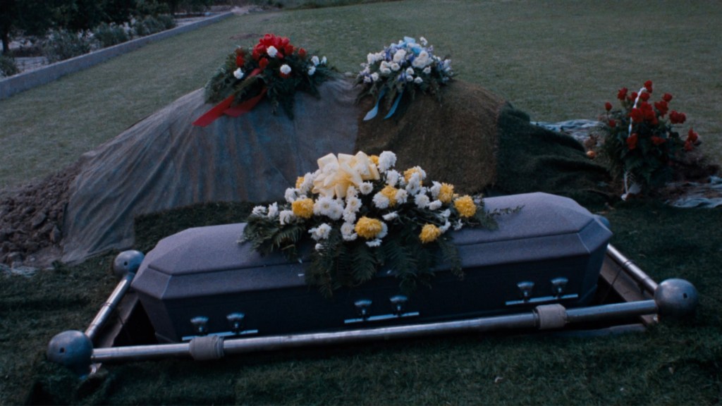 A grave site with a coffin adorned with various flowers, set in a grassy area with a mound of soil nearby.