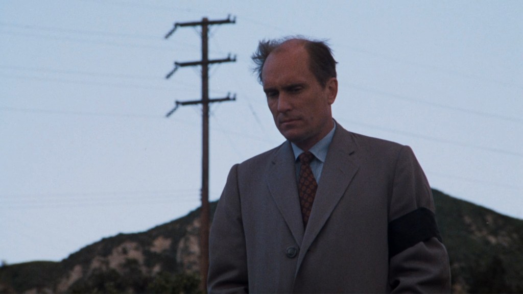 A man in a gray suit stands looking down, with a black armband, against a backdrop of telephone poles and a distant hillside.