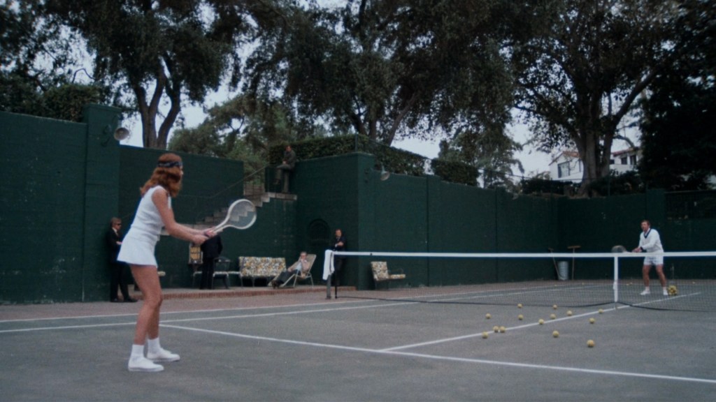 A woman in tennis attire prepares to hit a tennis ball on a court, while a man stands at the opposite end ready to receive. Surrounding the court are trees and spectators in the background.