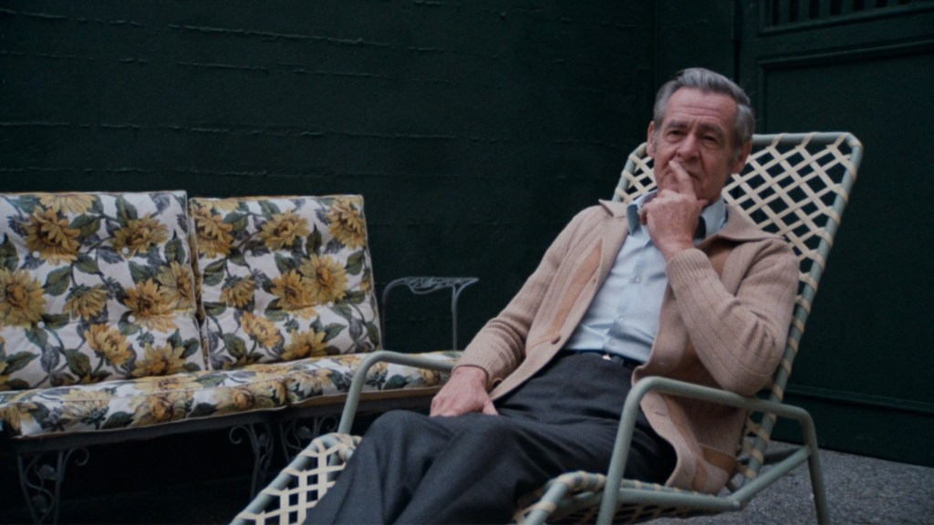 An older man sitting thoughtfully in a cushioned chair outdoors, surrounded by floral-patterned furniture against a dark green backdrop.