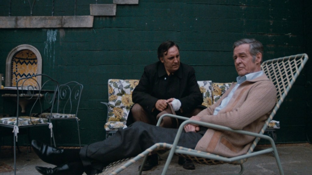 Two men sitting outdoors in a patio; one man in a reclining chair appears relaxed, while the other, standing with a bandaged hand, engages in conversation.