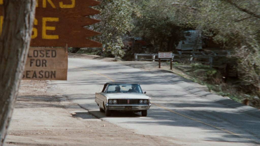 A vintage car driving on a winding road with a sign indicating it's closed for the season in the background.