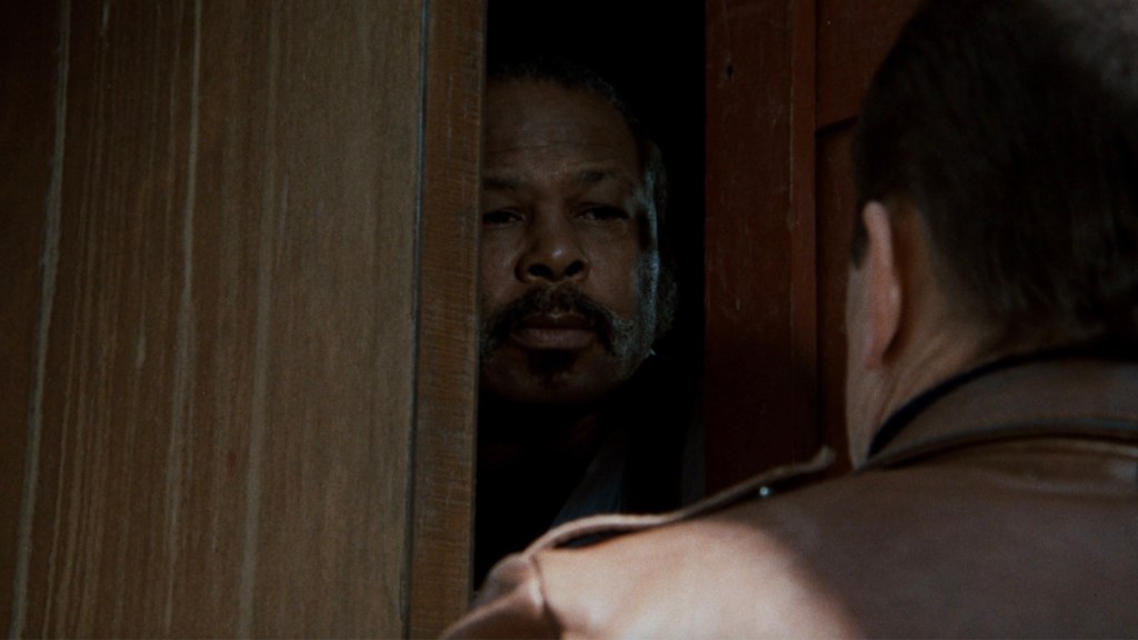 A man with facial hair peering through a partially open wooden door, gazing intently at another person off-camera.