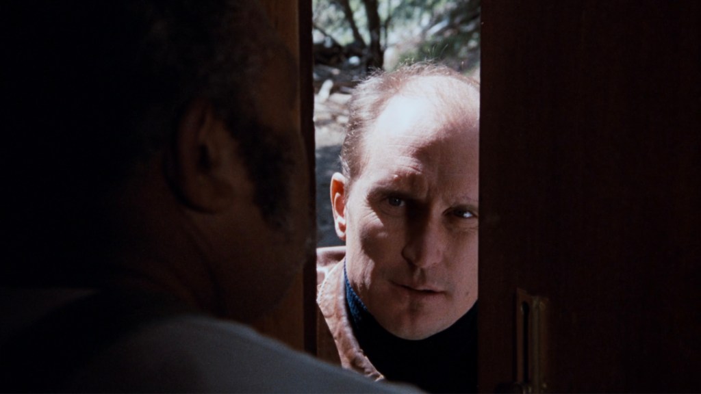 A tense moment at a door, showing a man peering through a partially opened door, while another man's face is visible from the side, suggesting a confrontation.