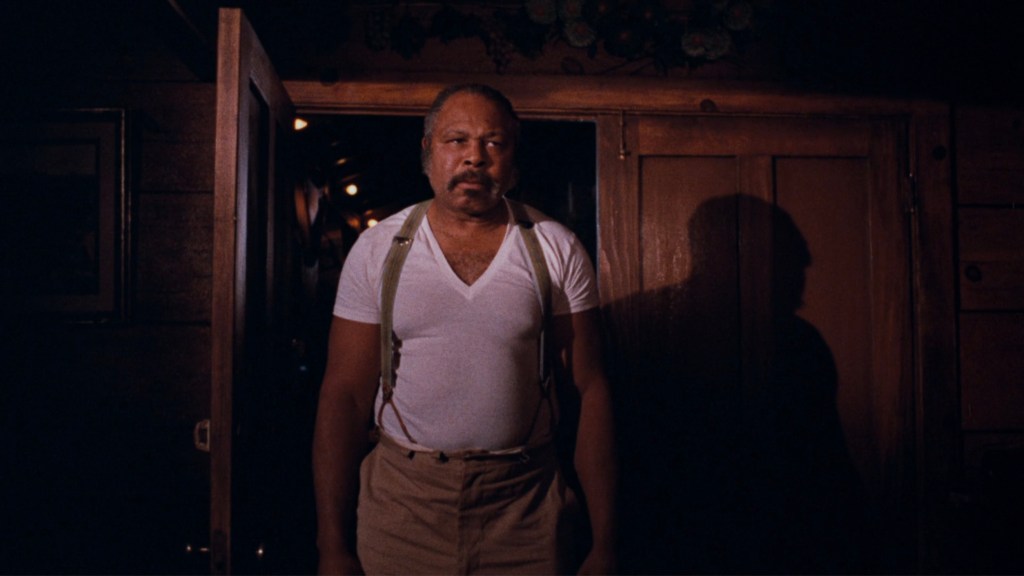 A man in a white tank top and beige trousers stands at an open door, partially illuminated, with a serious expression on his face and a shadow on the wall behind him.