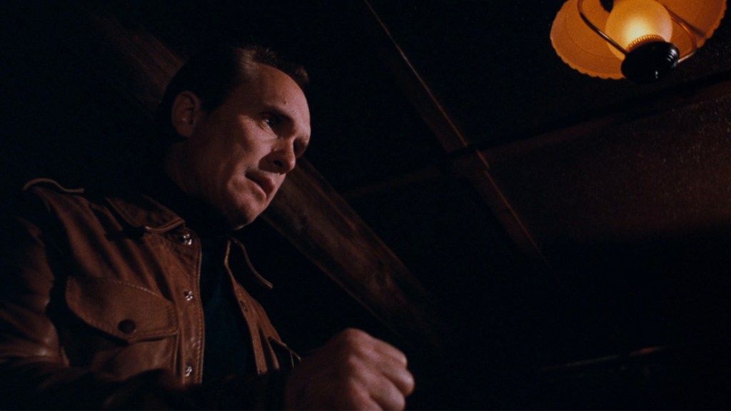 A man in a brown leather jacket stands under a dimly lit ceiling lamp, looking down with a serious expression.