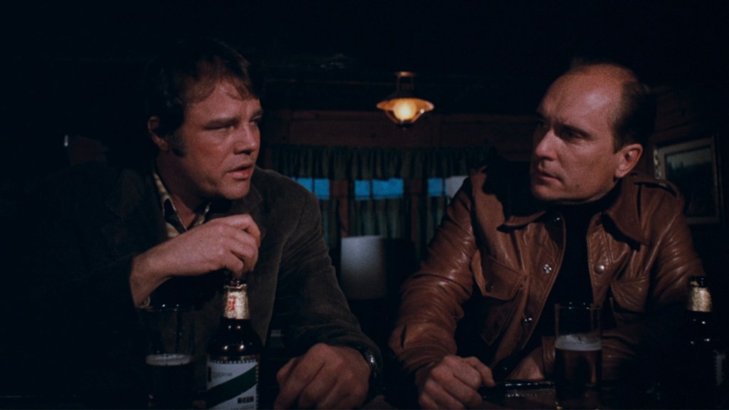Two men engaged in a serious conversation at a bar, with beer bottles on the table.