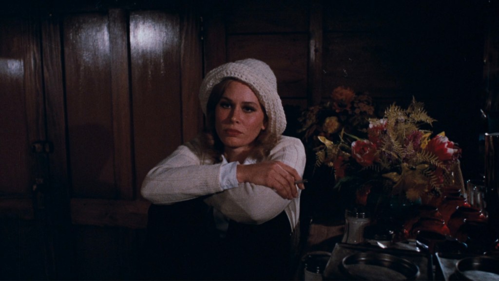 A woman sitting on the floor, wearing a white knitted hat and sweater, resting her chin on her knee while holding a cigarette. She is surrounded by a table with flowers and various objects in a dimly lit room.