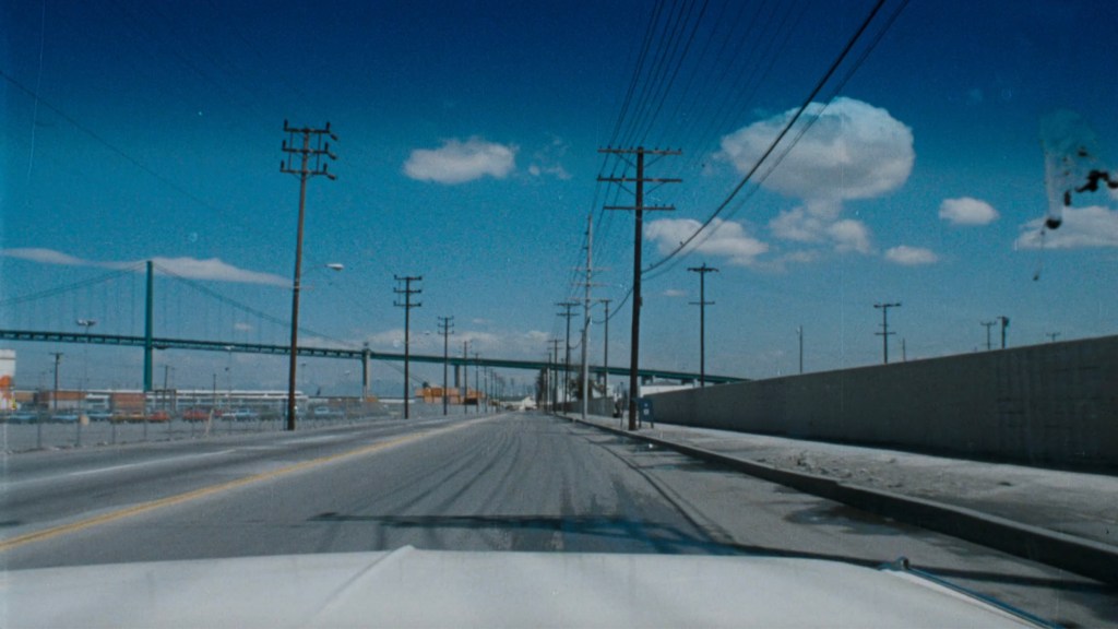 A view of a deserted road with power lines on both sides, leading towards a bridge in the background under a clear blue sky with scattered clouds.