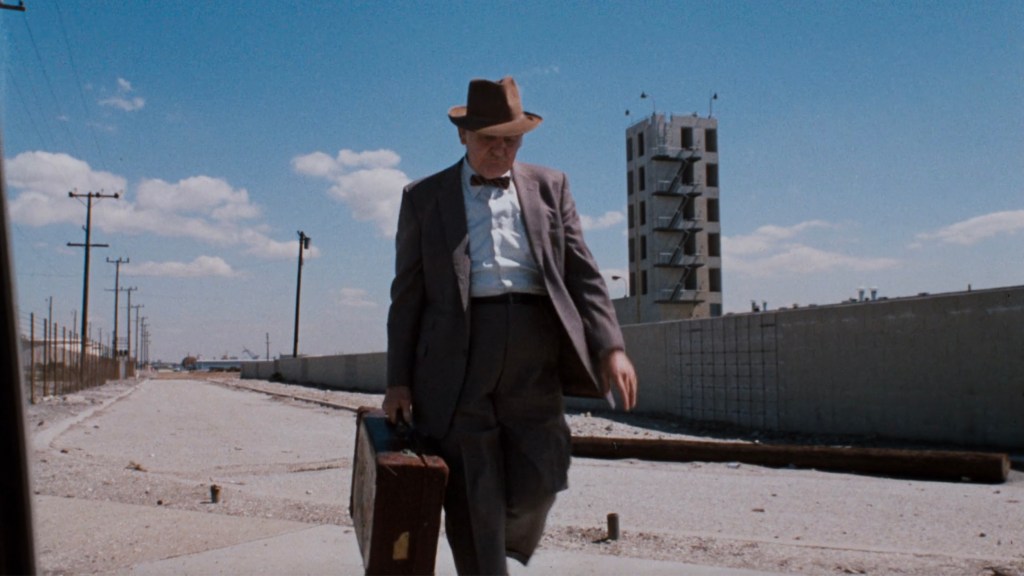 A man in a gray suit and hat walks along a desolate road carrying a briefcase, with a concrete wall and a tall structure in the background under a blue sky.