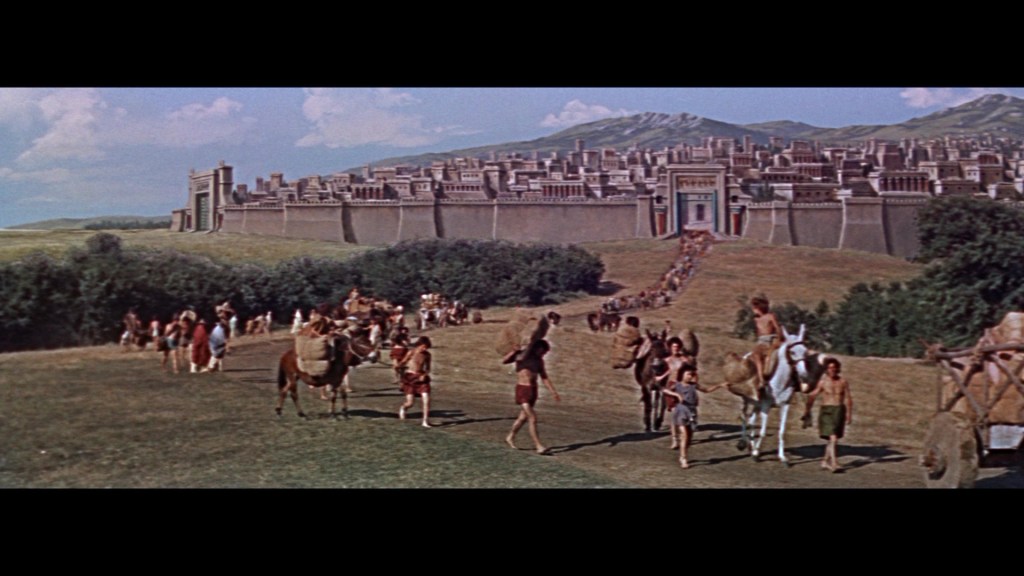 A historical scene depicting a large ancient city with towering walls in the background. People, including children and adults, are seen walking and carrying goods, with animals like horses and camels present.
