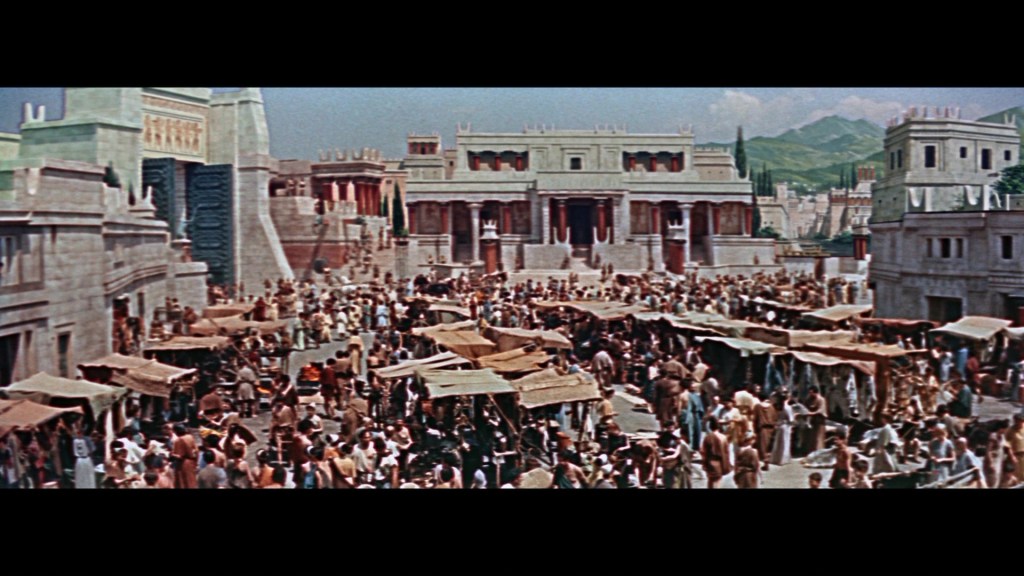 A bustling ancient marketplace filled with people, vendors, and tents set against a backdrop of grand stone buildings and mountains.