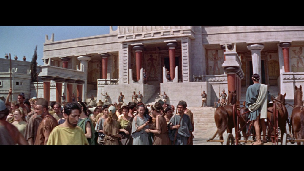 A crowded ancient marketplace with people in historical attire, featuring a grand stone building in the background adorned with columns and sculptures.