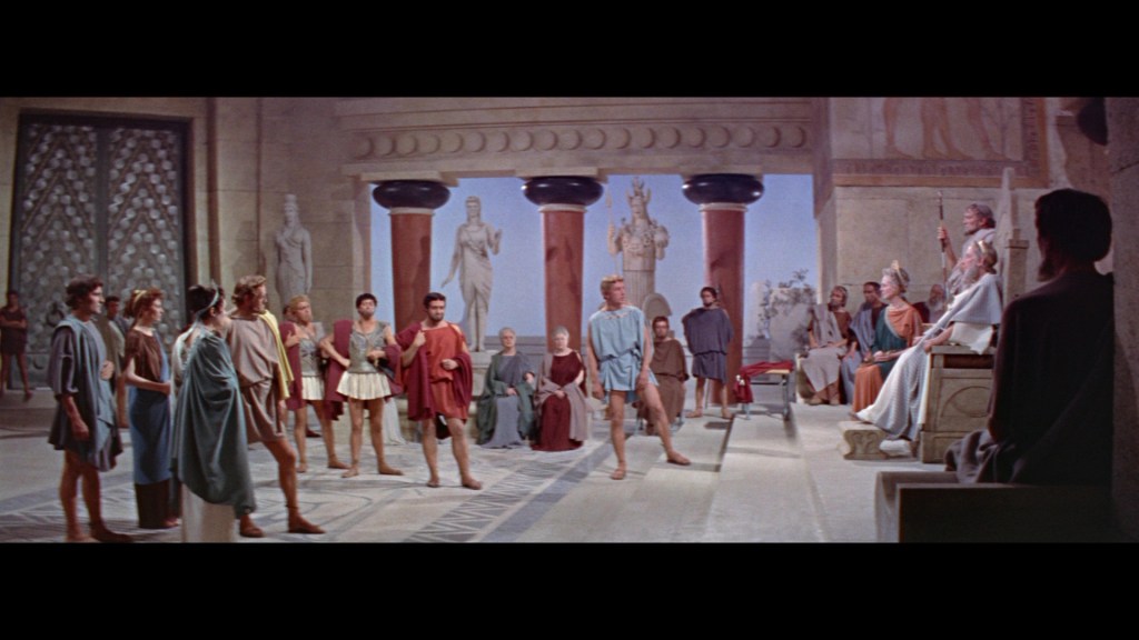 A scene depicting a group of ancient Greek figures in a large hall, with statues and architectural elements in the background. Some individuals are standing, while others are seated, creating a dramatic assembly atmosphere.