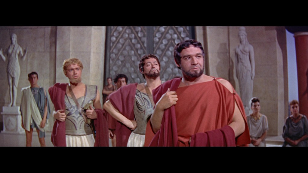 A group of men wearing ancient Roman togas and costumes in a grand hall with stone statues in the background.